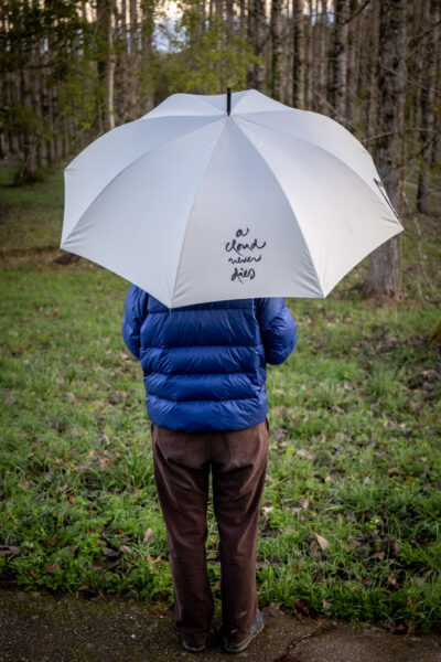Practitioner walking peacefully under Plum Village Calligraphy Umbrella showing A Cloud Never Dies zen calligraphy on sunny path; arapluie Calligraphie Plum Village sous la pluie pour marcher en pleine conscience